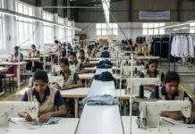 Sri Lanka Apparel Exports: The $8 Billion Challenge by 2030 Row of sewing stations in a garment factory with workers sewing fabric pieces at their machines, racks of clothes along the sides.
