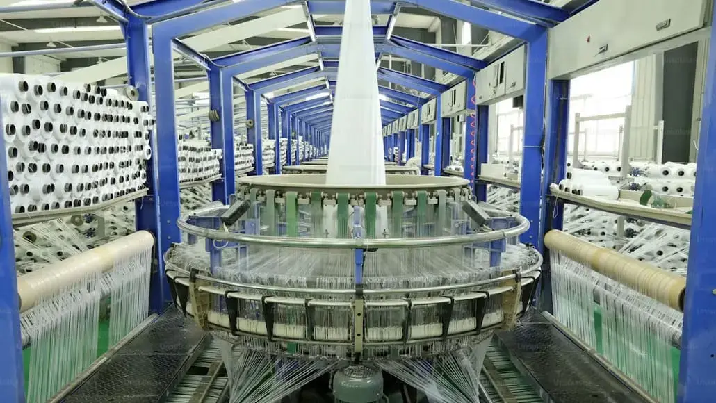 Circular knitting loom in a textile factory, blue steel frame with yarns feeding from spools on both sides.