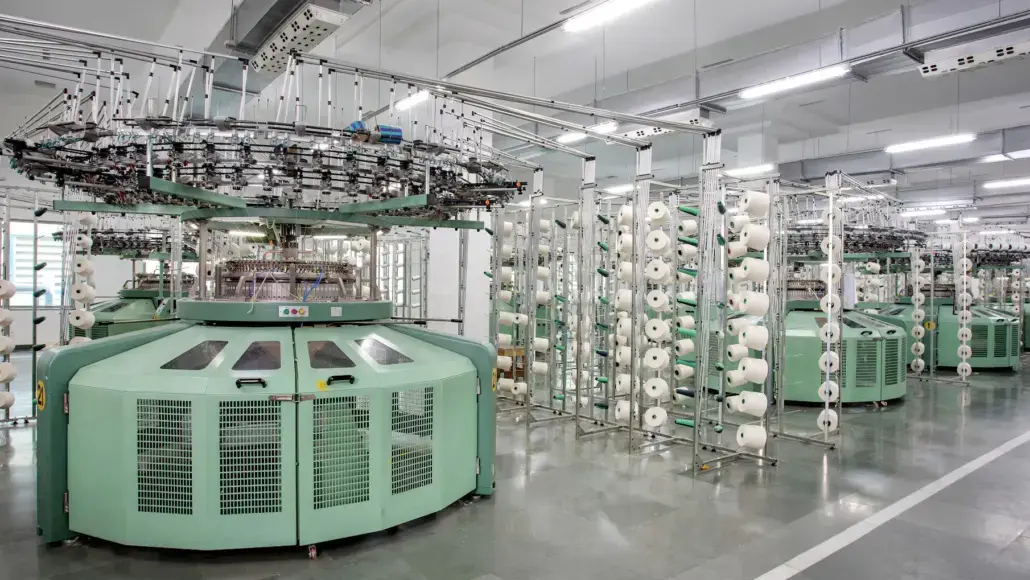 Green circular knitting machine in a modern textile factory, with multiple yarn spools and overhead threading infrastructure.