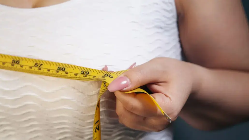 A person’s hands hold a yellow measuring tape around their waist, measuring for fit, against a white textured dress.